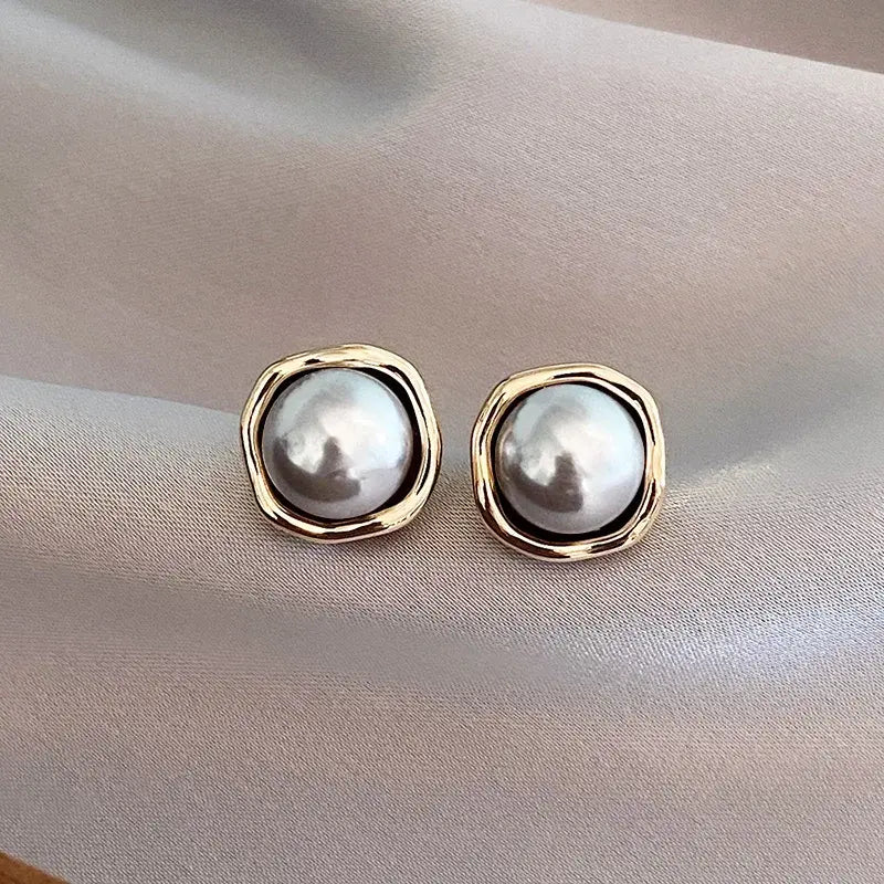 A pair of black pearl stud earrings with a gold-tone rim on gray satin